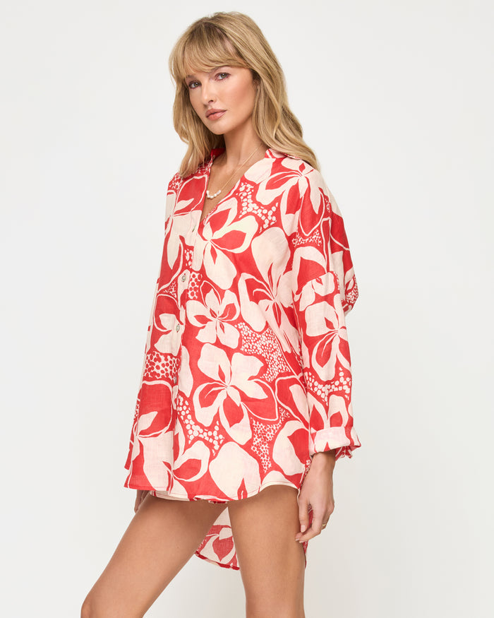 Three-quarter side view of a model wearing the LSPACE Rio Tunic in hibiscus bloom, a long-sleeve collared button-front shirt in the red and cream hibiscus print styled over matching shorts.