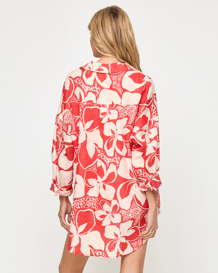Back view of a model wearing the LSPACE Rio Tunic in hibiscus bloom, showing the full back coverage of the long-sleeve button-front shirt in the red and cream hibiscus floral print.