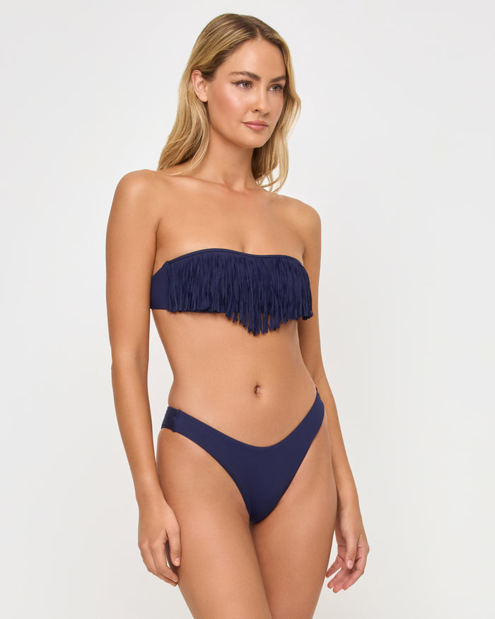 Three-quarter front view of a model wearing the LSPACE Rocky Bikini Bottom in Sensual Solid Midnight Blue navy, paired with a navy fringe bandeau Dixie Bikini Top.