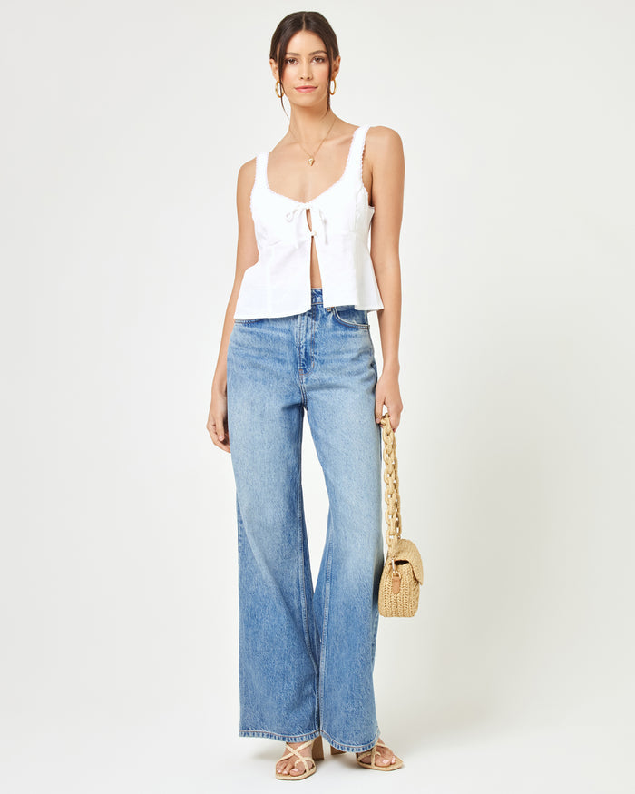 Front view of a model wearing the LSPACE Rosa Top in cream, a sleeveless top with a scoop neckline, tie-front detail, and flared peplum hem, styled with wide-leg blue jeans and a straw bag.