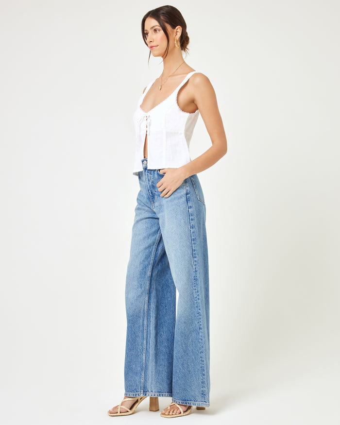 Side view of a model wearing the LSPACE Rosa Top in cream, showing the tie-front detail and flared peplum silhouette, styled with wide-leg blue jeans and strappy heels.