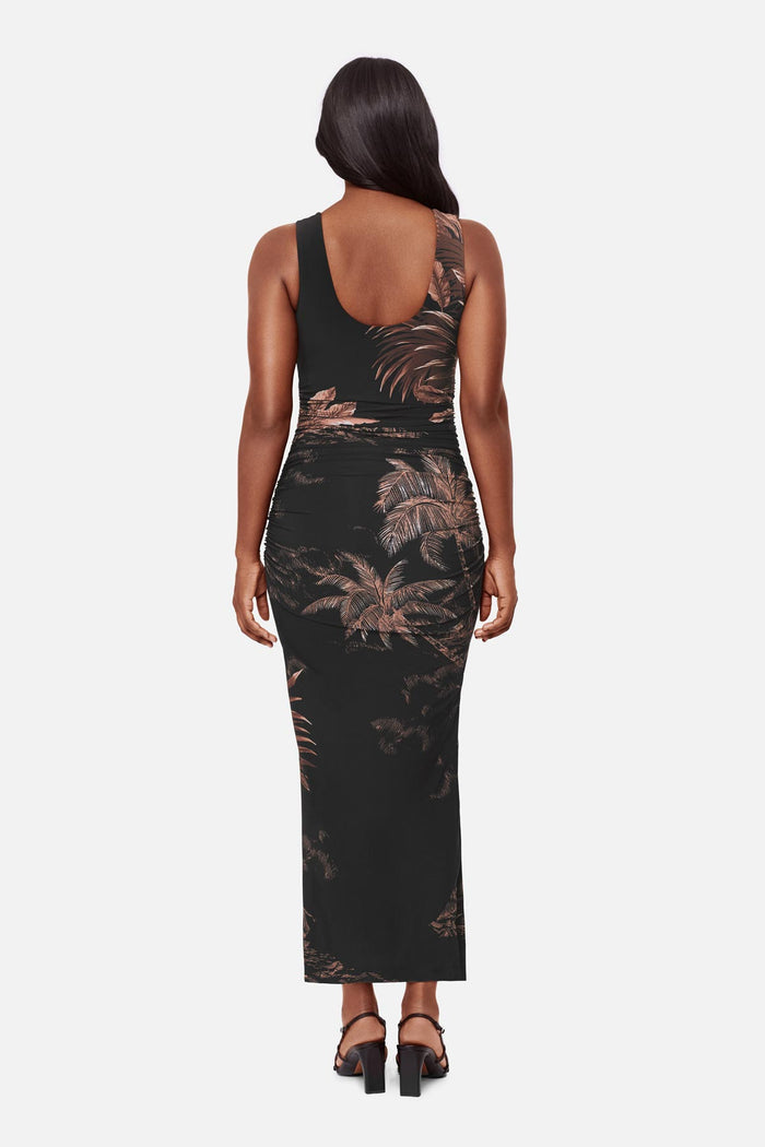 Woman turned away wearing a sleeveless full-length dress with a brown and shite detailed palm tree print