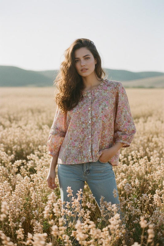 May include: A woman wearing a floral print blouse with a round neckline and button-up front, paired with blue jeans. The blouse features a pink and purple floral pattern. The background is a field of wildflowers.