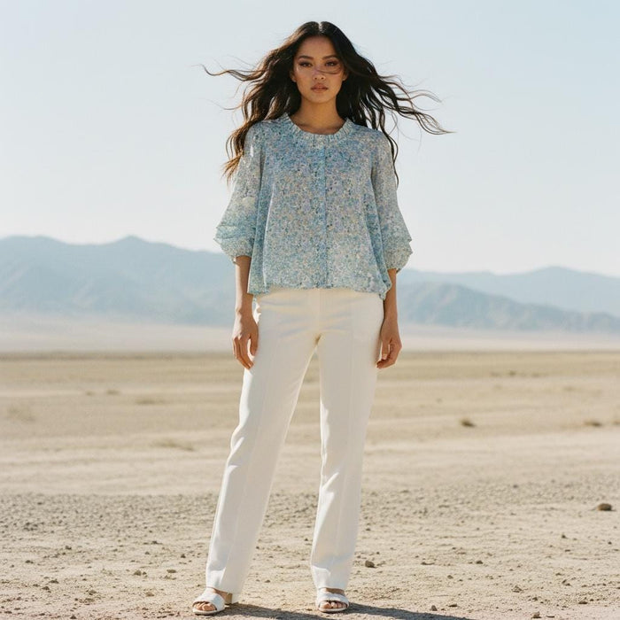 May include: A woman wearing a light blue floral print blouse with three-quarter sleeves and white pants. The blouse has a round neckline and a button-down front. The pants are straight-legged and the woman is wearing white sandals. The background is a desert landscape.