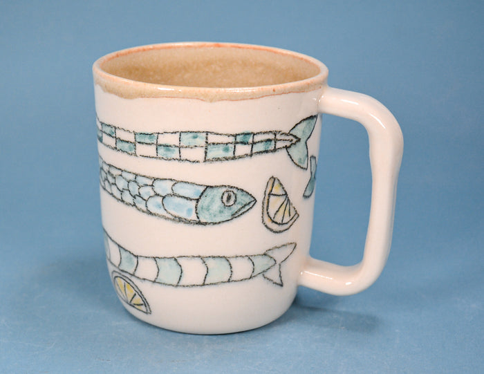 A white ceramic mug with a handle, decorated with hand-painted fish and lemon slice motifs in light blue and green, set against a solid blue background.