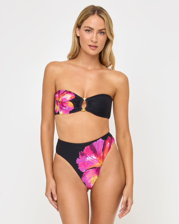 Front view of a model wearing the LSPACE after midnight hibiscus Savannah Bikini Bottom, a high-waist style with hibiscus floral print in pink and orange on a dark background, paired with a matching strapless bandeau Jasper Bikini Top with gold O-ring detail.