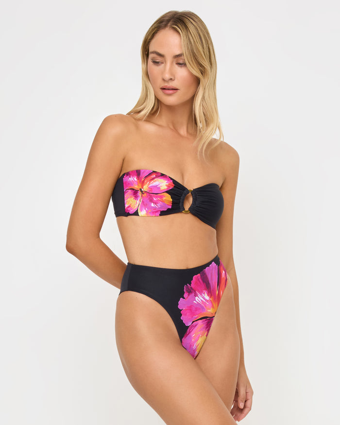 Three-quarter side view of a model wearing the LSPACE after midnight hibiscus Savannah Bikini Bottom, a high-waist style with hibiscus print on the sides, paired with a matching strapless bandeau Jasper Bikini Top with gold O-ring.