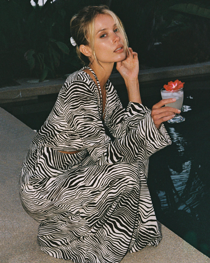 Editorial view of a model crouching poolside wearing the LSPACE Say When Maxi Dress in animal, a long-sleeve maxi dress with a front cutout and black and white zebra print.