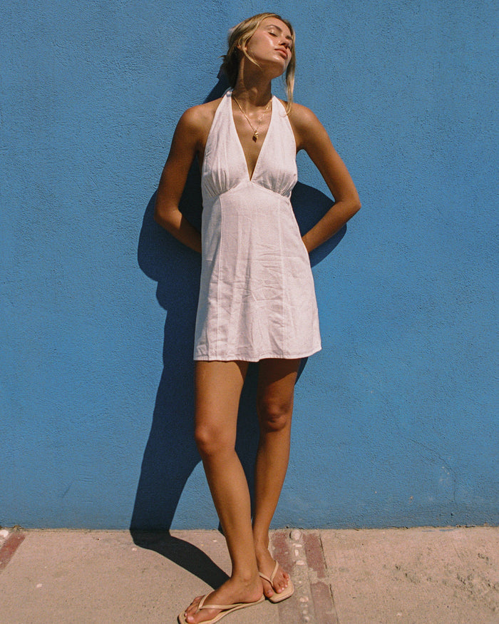 Front view of a model wearing the LSPACE Seabreeze Dress in cream, a lightweight halter mini dress with a deep V-neckline and ruched empire waist, styled with flat sandals against a bright blue wall.