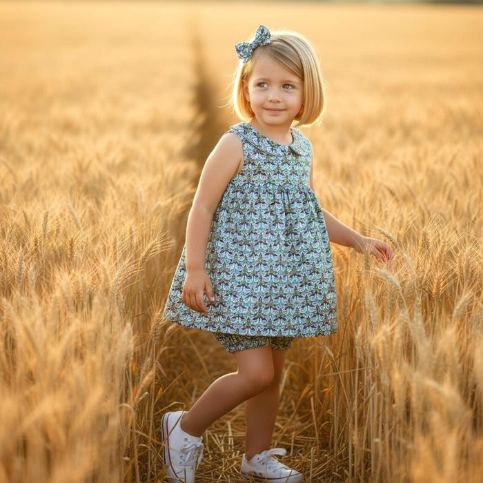 May include: A young girl in a blue and green patterned dress and matching bow stands in a wheat field. She wears white sneakers. The dress has a Peter Pan collar and a floral pattern. The background is a golden wheat field.