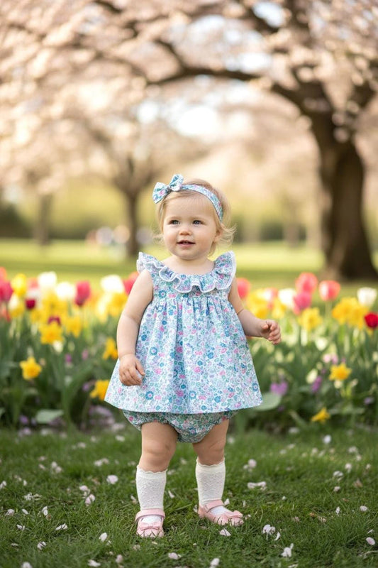 May include: A baby wearing a blue floral print dress, bloomers, and matching headband. The dress has ruffled sleeves and a white trim. The baby is also wearing white socks and pink shoes, standing in a field of flowers.