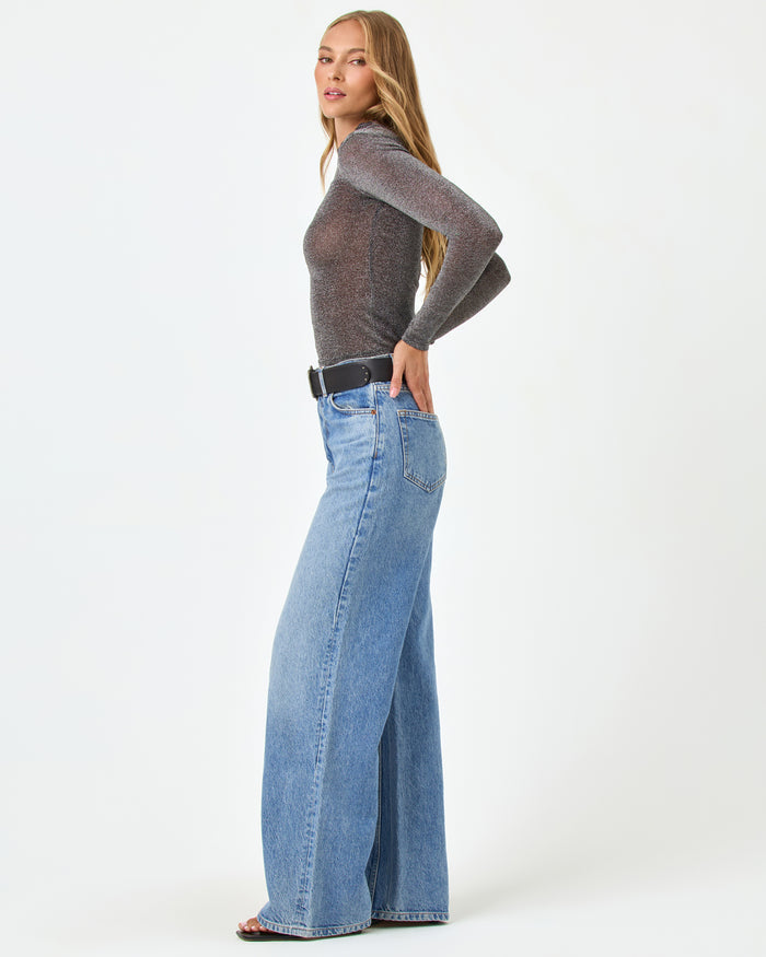 Side view of a model wearing the LSPACE Shimmer Long Sleeve in black, showing the sheer metallic sparkle fabric and fitted silhouette, styled with wide-leg jeans and a black belt.
