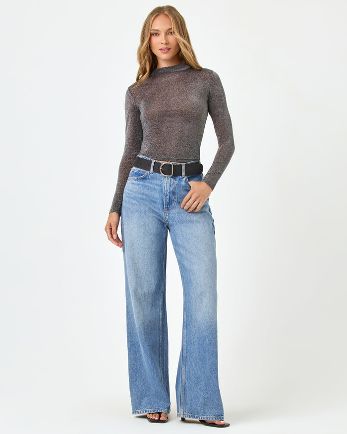 Front view of a model wearing the LSPACE Shimmer Long Sleeve in black, a sheer metallic long-sleeve top with mock neckline and cropped hem, styled with wide-leg jeans and a black belt.