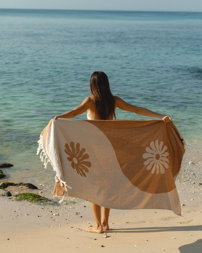 Model on the beach holding the Slowtide Botanic Balance Turkish Towel in brush open behind her back, facing away toward the ocean, displaying the tan and cream botanical pattern.