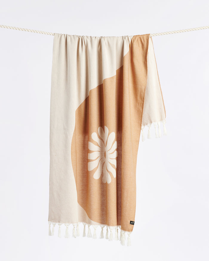 The Slowtide Botanic Balance Turkish Towel in brush hanging on a rope, showing the yin-yang botanical pattern with daisy motif in tan and cream with fringe.