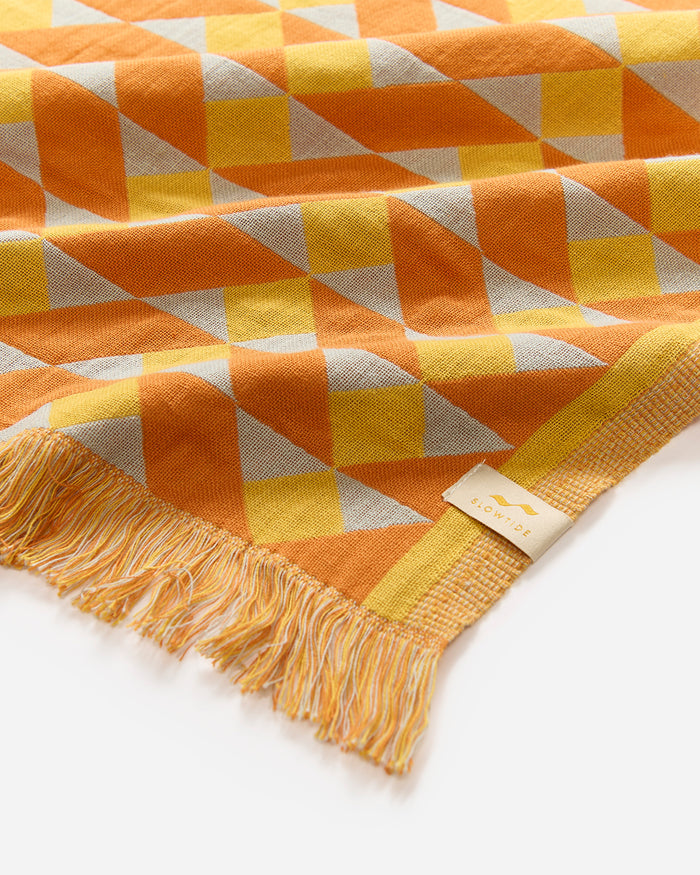 Close-up corner detail of the Slowtide Daydream Driftweave Turkish Towel in curry, showing the geometric triangle pattern in orange, yellow and grey with fringe and the Slowtide woven label.