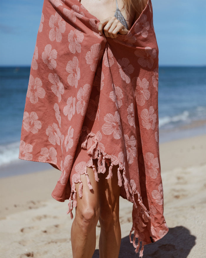 Model on the beach wrapped in the Slowtide Hau Turkish Towel in warm earth, displaying the hibiscus pattern and matching terracotta fringe.