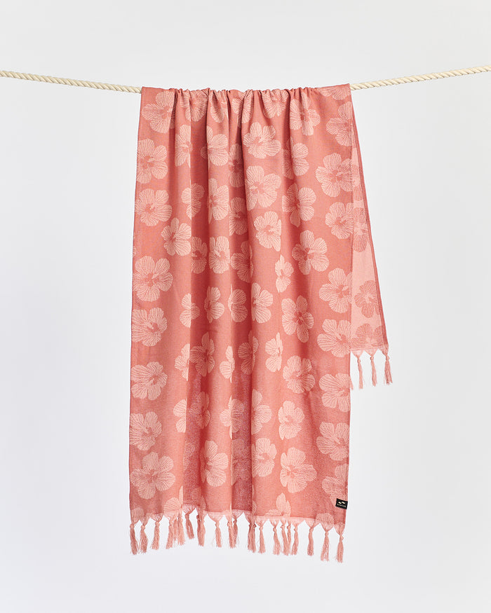 The Slowtide Hau Turkish Towel in warm earth hanging on a rope, displaying the all-over hibiscus pattern in terracotta and blush with matching fringe.