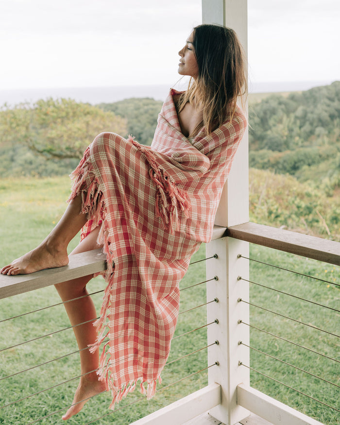 Model wrapped in the Slowtide Palaka Lightweight Beach Blanket in guava while seated on a balcony railing overlooking greenery.