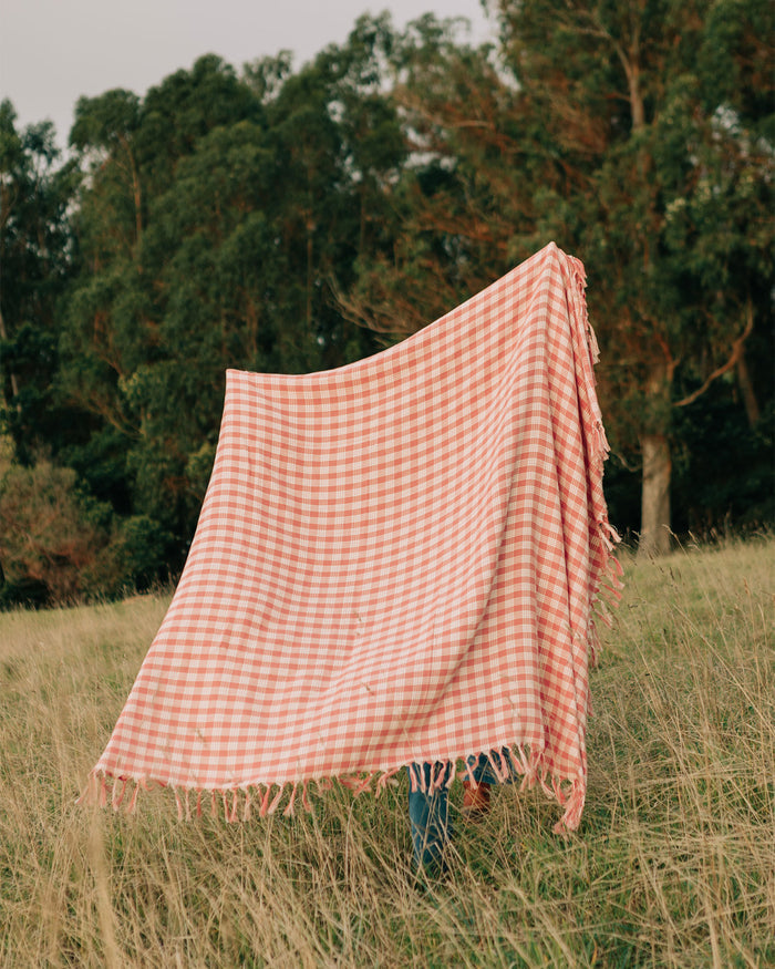The Slowtide Palaka Lightweight Beach Blanket in guava held up outdoors in a field with trees in the background.
