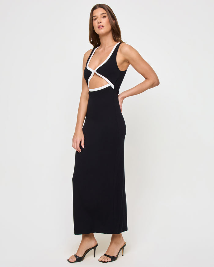 Side view of a model wearing the LSPACE Steph Dress in black-cream, showing the form-fitting maxi silhouette with a side slit and crossover cut-out bodice with cream trim, styled with heeled mule sandals.