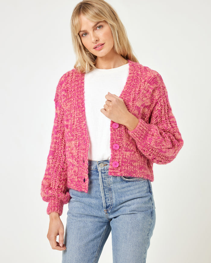 Front view of a model wearing the LSPACE Strawberry Fields Cardigan in strawberry marl, a pink cable-knit button-front V-neck cardigan with pink buttons, styled open over a white tee and jeans with sneakers on a white background.