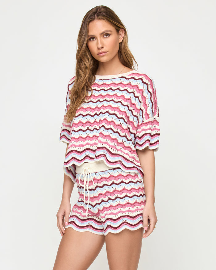 Front view of a model wearing the LSPACE sand in hand Sunchaser Short, crochet knit shorts with a multicolor wavy stripe pattern in pink, blue, and white, styled with the matching Sunchaser Top, on a neutral studio background.