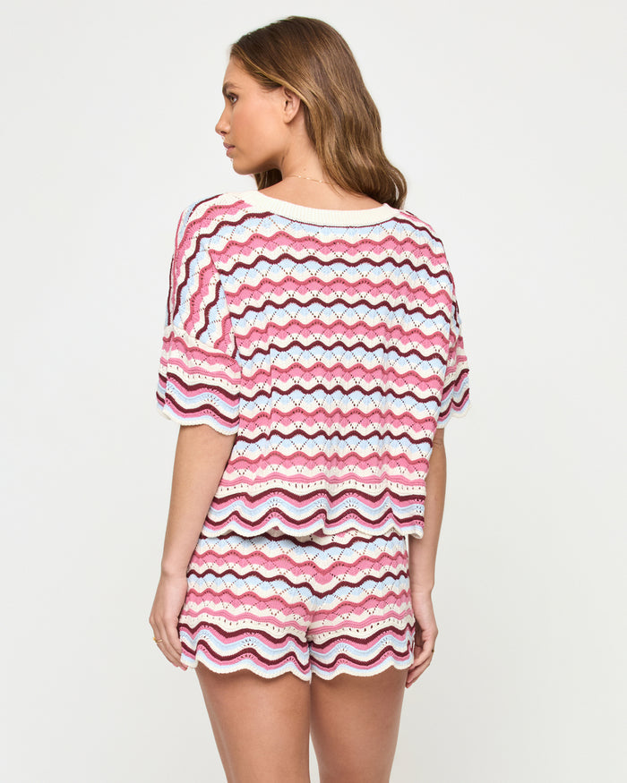 Back view of a model wearing the LSPACE sand in hand Sunchaser Short, crochet knit shorts with a multicolor wavy stripe pattern in pink, blue, and white, styled with the matching Sunchaser Top, on a neutral studio background.