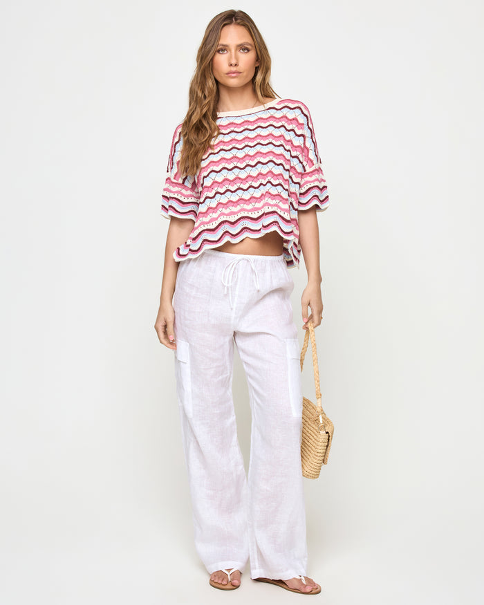 Front view of a model wearing the LSPACE sand in hand Sunchaser Top, an oversized crochet knit short-sleeve crop top with a multicolor wavy stripe pattern in pink, blue, and white, styled with white wide-leg pants, on a neutral studio background.