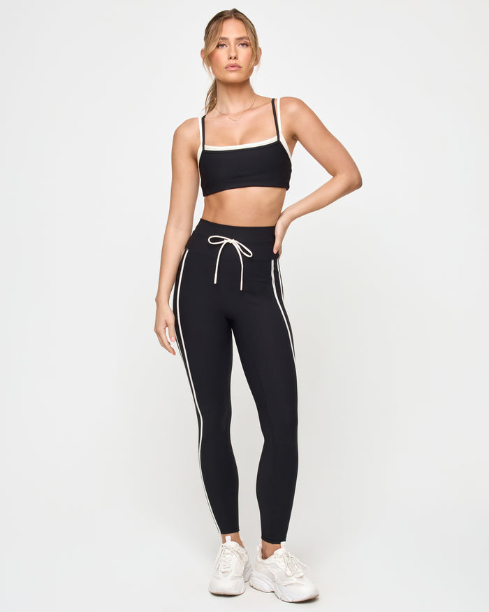 Front view of the LSPACE Sutton Pant in black-cream on a model against a light background, showing the high-rise athletic pants with a cream side stripe and drawstring waist, paired with a black-cream sports bra.