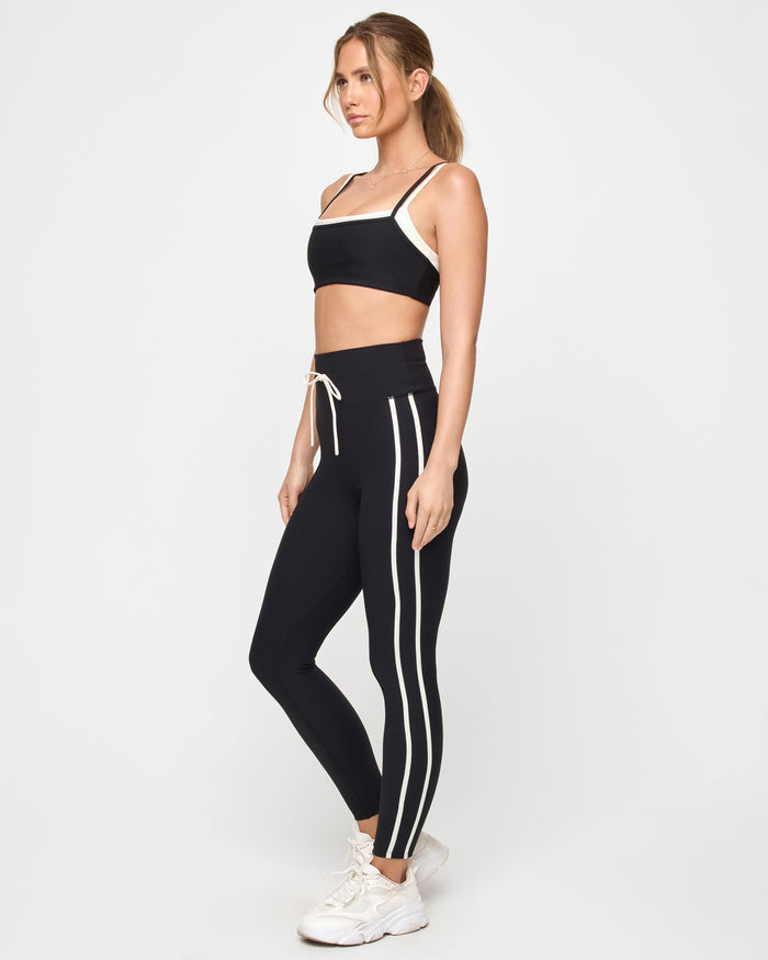 Side view of the LSPACE Sutton Pant in black-cream on a model against a light background, showing the cream side stripe along the back of the high-rise athletic pants, paired with a black-cream sports bra.
