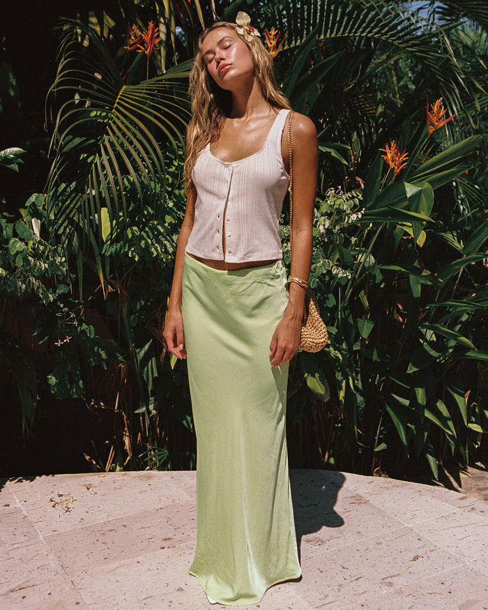 Front view of a model wearing the LSPACE Sweet As Honey Skirt in honeydew, a satin maxi skirt with a bias-cut silhouette, styled in a tropical garden setting.