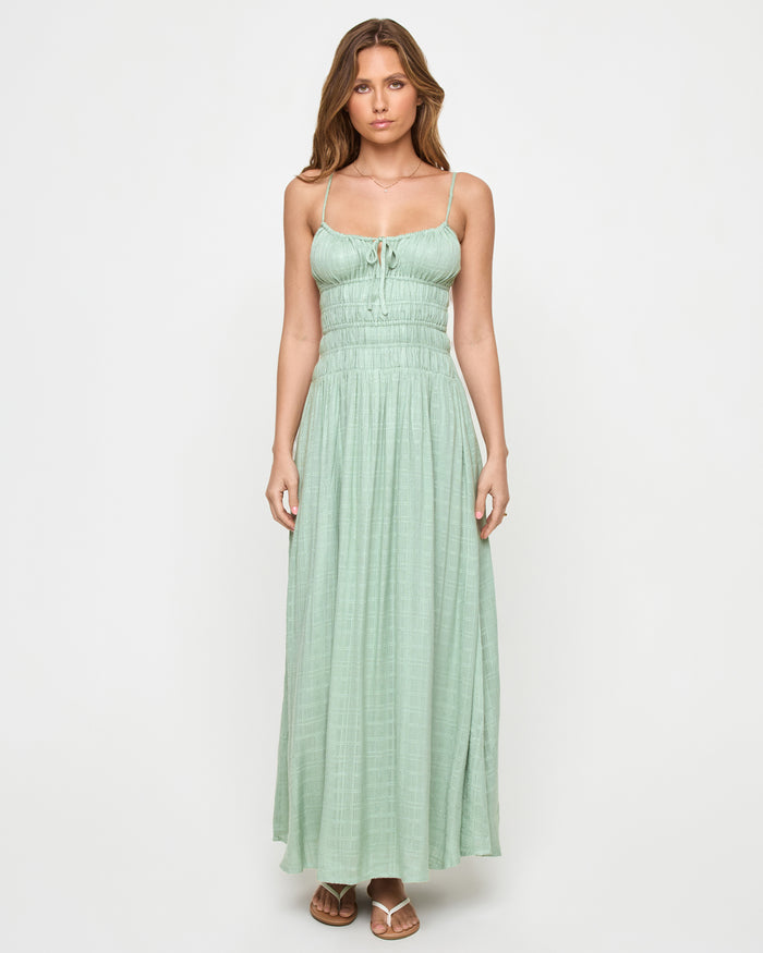 Front view of a model wearing the LSPACE Tala Dress in celadon, a spaghetti-strap maxi dress with a front tie detail, smocked empire waist, and flowing skirt in a sage green color.