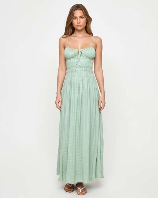Front view of a model wearing the LSPACE Tala Dress in celadon, a spaghetti-strap maxi dress with a front tie detail, smocked empire waist, and flowing skirt in a sage green color.
