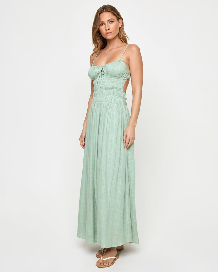 Three-quarter front view of a model wearing the LSPACE Tala Dress in celadon, showing the spaghetti straps, front tie bodice, side waist cutout, and flowing maxi skirt in sage green.
