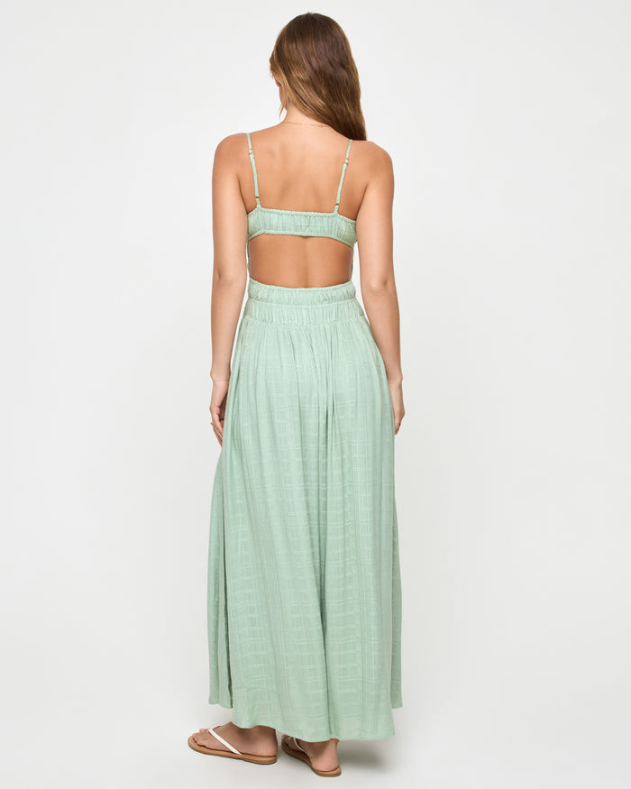 Back view of a model wearing the LSPACE Tala Dress in celadon, showing the open back with spaghetti straps and an open back cutout above the smocked waist in sage green.