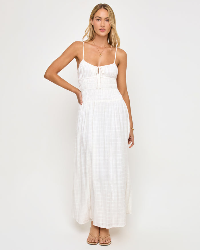 Front view of a model wearing the LSPACE Tala Dress in cream, a full-length maxi dress in an off-white textured fabric with thin spaghetti straps, a smocked bodice with a front center tie, and a flowing skirt.