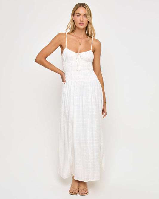 Front view of a model wearing the LSPACE Tala Dress in cream, a full-length maxi dress in an off-white textured fabric with thin spaghetti straps, a smocked bodice with a front center tie, and a flowing skirt.