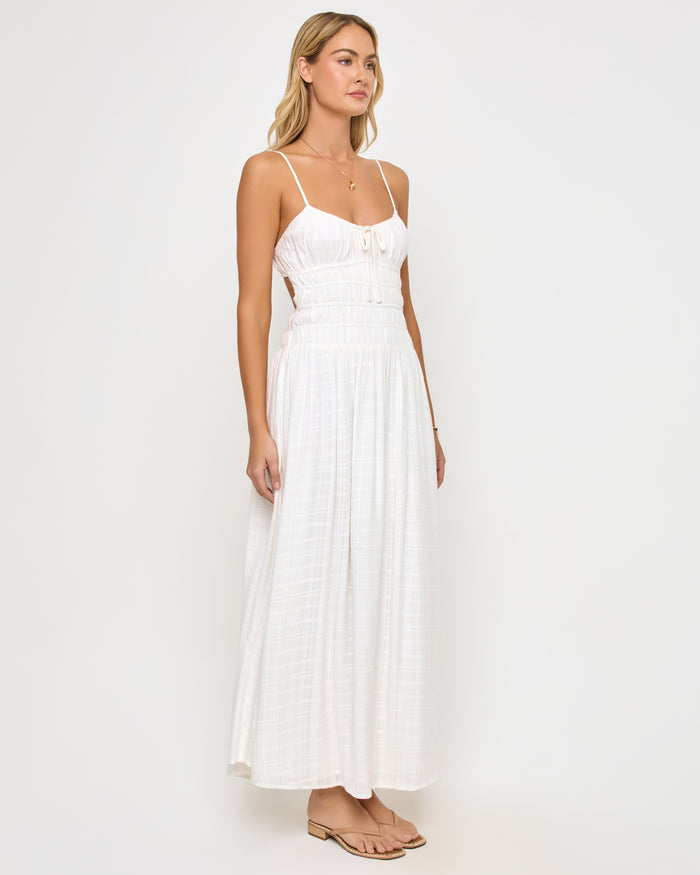 Angled view of a model wearing the LSPACE Tala Dress in cream, showing the open-back cutout design and the flowing off-white textured maxi skirt from the side.