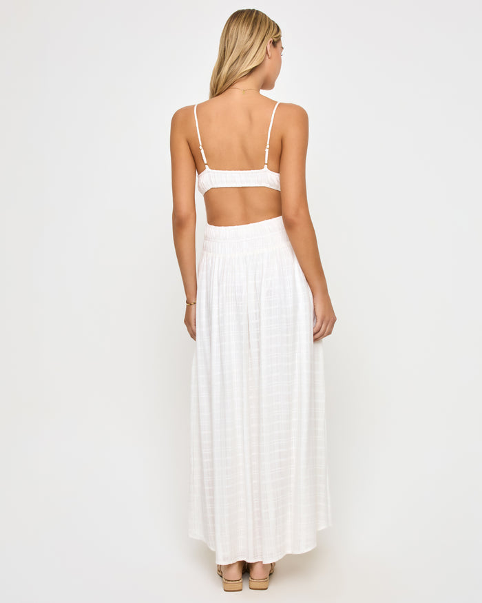Back view of a model wearing the LSPACE Tala Dress in cream, showing the wide open-back cutout between the crop-style bodice and smocked waistband of the off-white maxi dress.