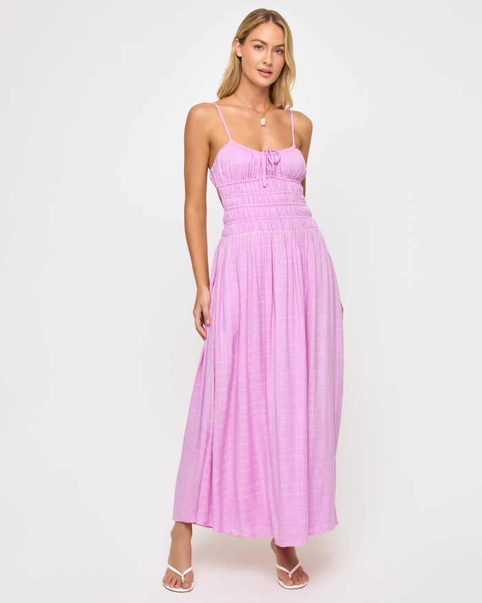 Front view of a model wearing the LSPACE Tala Dress in light iris, a full-length maxi dress in a soft lavender hue with thin spaghetti straps, a smocked bodice with a front center tie, and a flowing skirt.