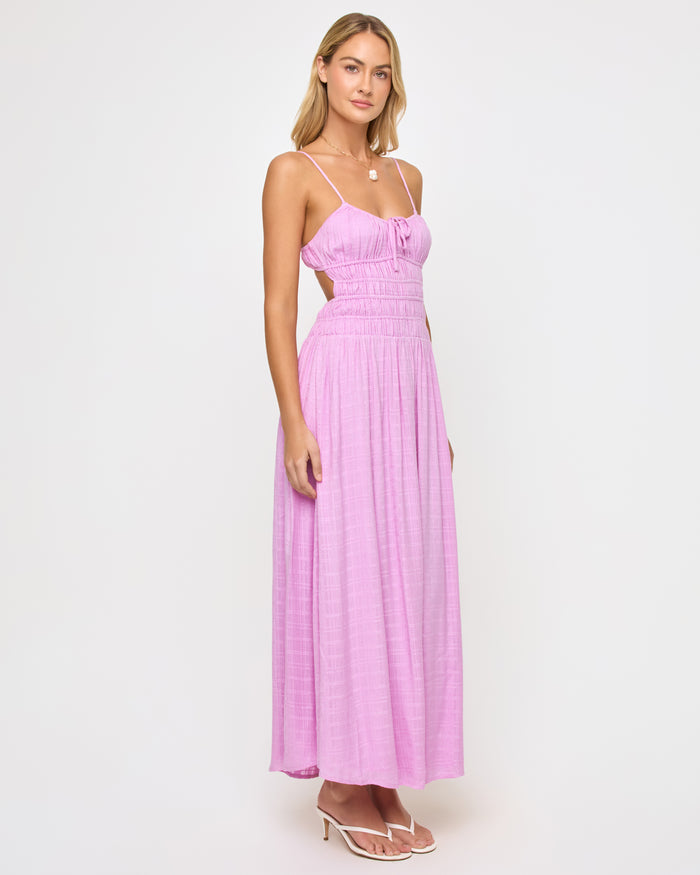 Angled view of a model wearing the LSPACE Tala Dress in light iris, showing the open-back cutout design with spaghetti straps and the flowing lavender maxi skirt from the side.