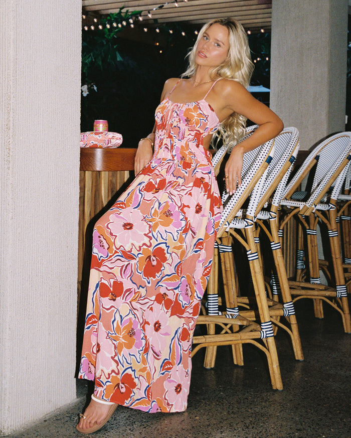 Campaign view of a model wearing the LSPACE Tala Dress in lost in the blooms, a maxi dress in a vibrant retro floral print with pink, orange, red, and blue blooms, photographed in an indoor bar setting with rattan chairs and string lights.