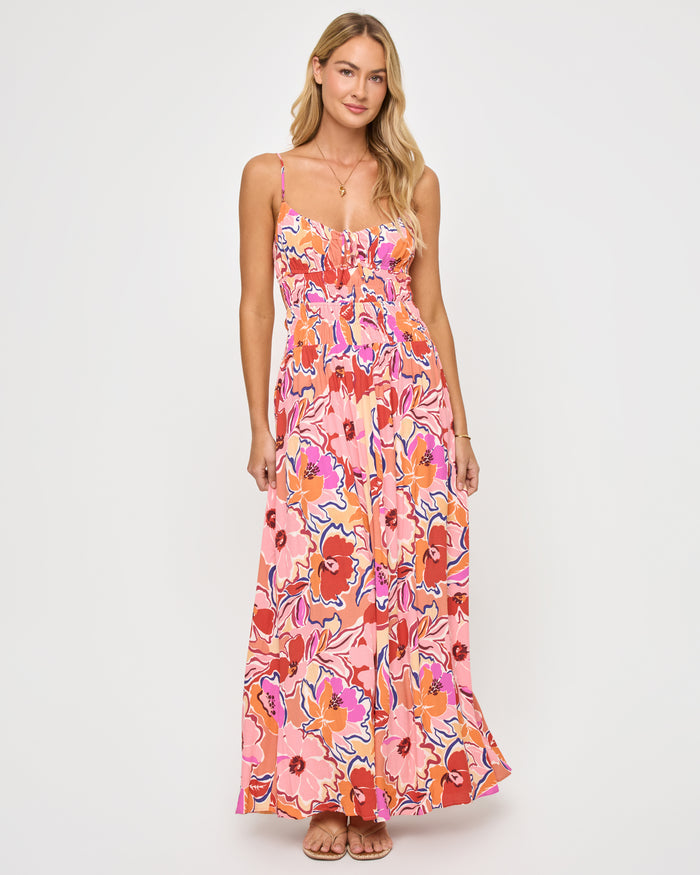Front view of a model wearing the LSPACE Tala Dress in lost in the blooms, a full-length maxi dress in a retro floral print with pink, orange, red, and blue blooms, featuring thin spaghetti straps, a smocked bodice, and a flowing skirt.