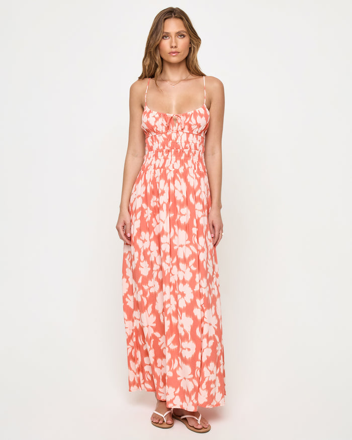 Front view of a model wearing the LSPACE Tala Dress in vine vignette, a spaghetti-strap maxi dress with a front tie detail, smocked empire waist, and coral and white floral print.