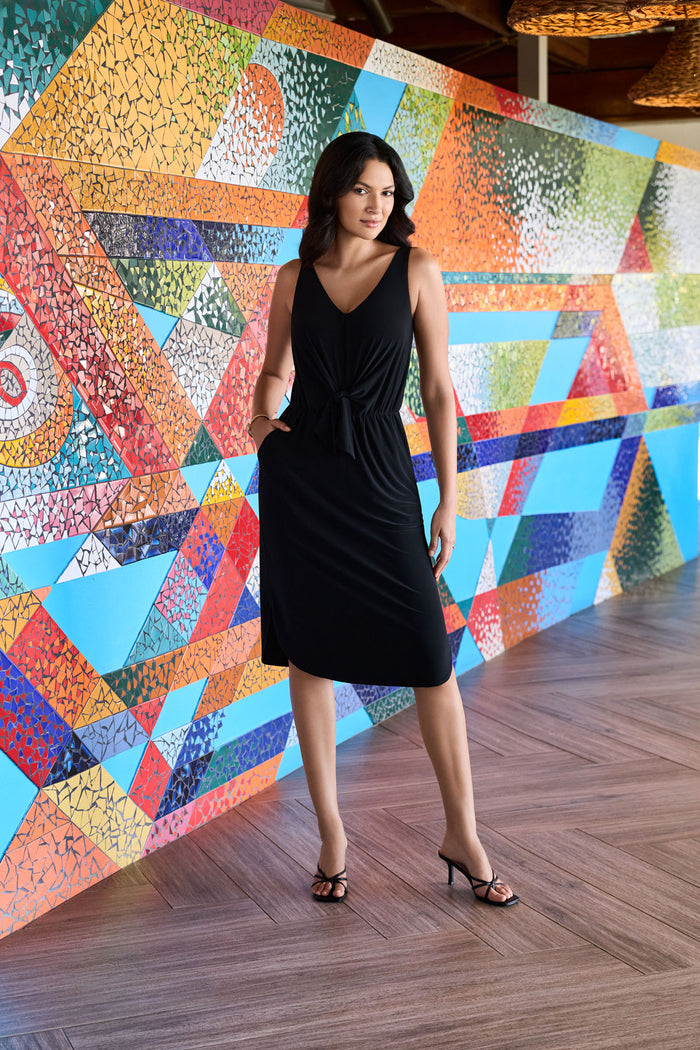 Woman next to bright colored wall wearing a black sleeveless mid length dress