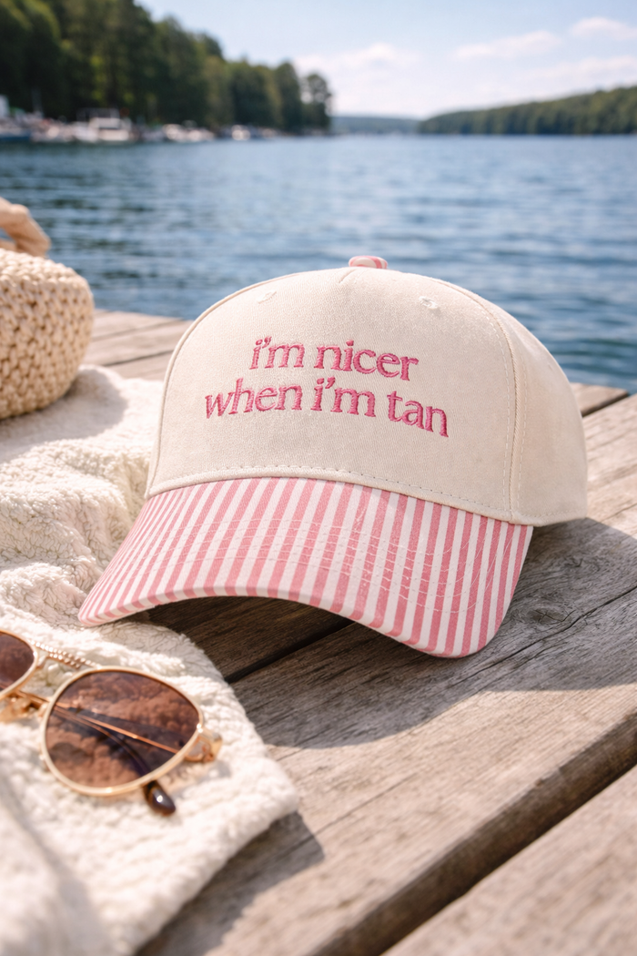 Beige cap with pink striped brim and 'I'm nicer when I'm tan' text, sunglasses, and towel on a wooden dock by water.