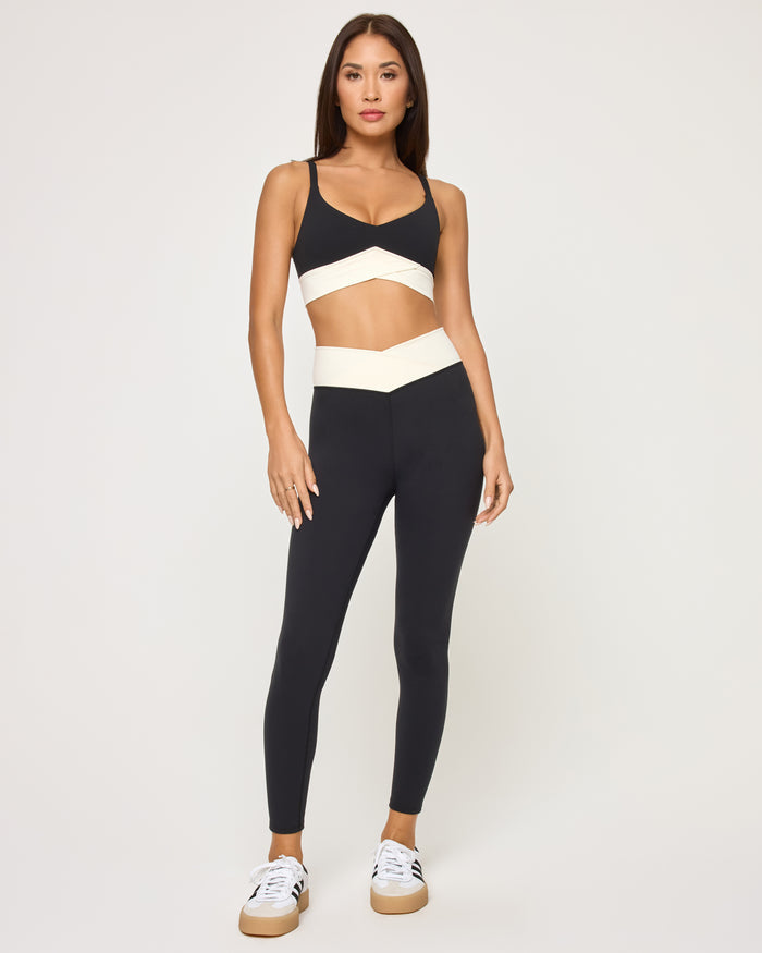 Front view of the LSPACE Unmatched Legging in black-cream on a model against a white background, paired with the matching black triangle sports bra with cream crossover band, showing the cream crossover waistband detail of the full-length leggings.