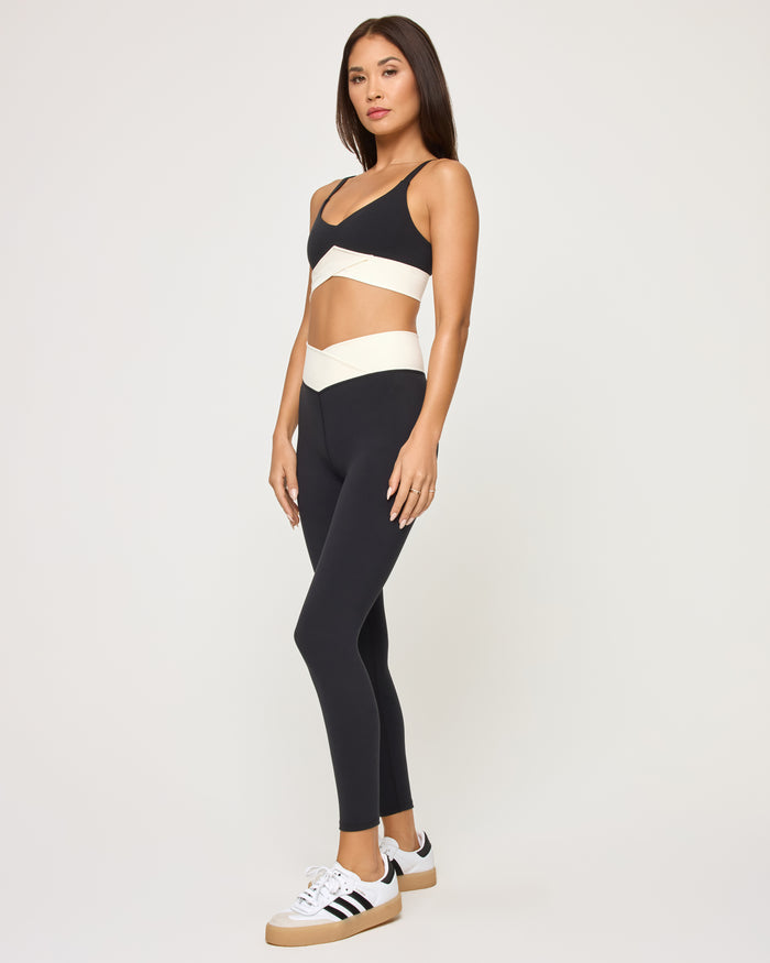 Side view of the LSPACE Unmatched Legging in black-cream on a model against a white background, paired with the matching black crossover sports bra with cream band detail, and black and white sneakers.