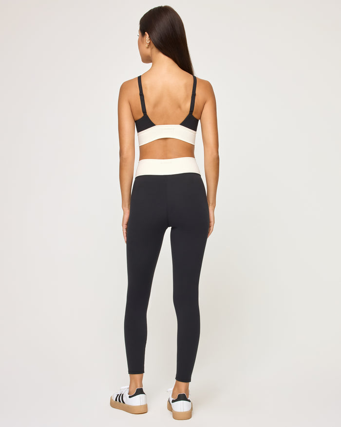 Back view of the LSPACE Unmatched Legging in black-cream on a model against a white background, showing the cream crossover waistband detail and the open back of the matching black sports bra, with black and white sneakers.